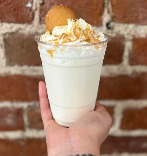 A hand holding a cup of creamy dessert topped with whipped cream, a cookie, and drizzled caramel against a brick wall.