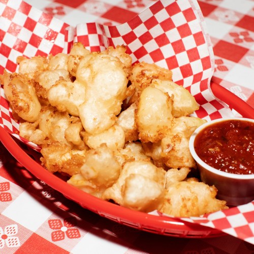 A red basket filled with golden fried cauliflower bites and a small cup of dipping sauce on a checkered tablecloth.