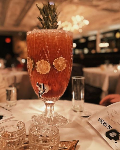 A large, ornate glass filled with a tropical drink, garnished with a pineapple, sits on a table in a restaurant.