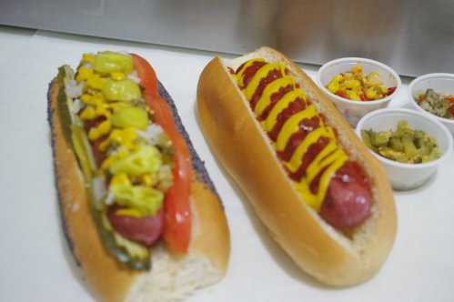 Two hot dogs in buns, one topped with mustard, ketchup, and relish, the other with onions, peppers, and yellow mustard.