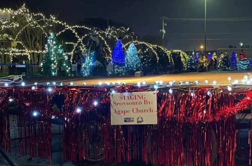 A festive display of colorful Christmas trees and lights at night, with a sign reading "Spacing Sponsored By Agape Church."