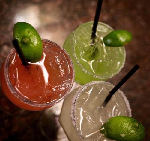 Three colorful cocktails with lime wedges, served in glasses with ice and straws on a dark countertop.