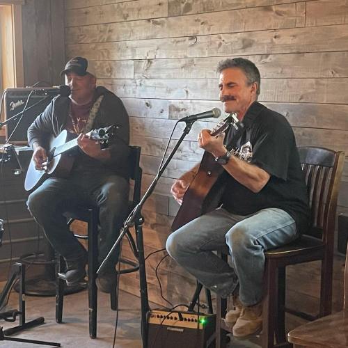 Two musicians perform live in a cozy venue, one on acoustic guitar and the other on vocals, both seated.