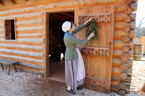 A woman in historical attire hangs greenery on a wooden door of a log cabin.