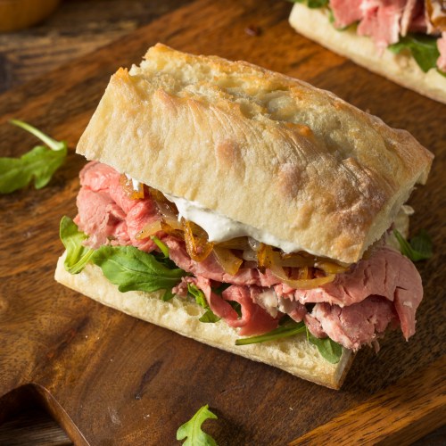 A hearty sandwich with layers of roast beef, onions, and greens on a crusty bread, served on a wooden board.