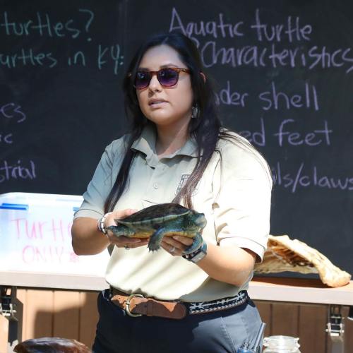 A woman in a polo shirt holds a turtle, standing in front of a chalkboard with turtle information.
