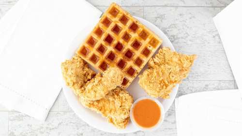 A plate with crispy fried chicken, a golden waffle, and a small cup of sauce on a light background.