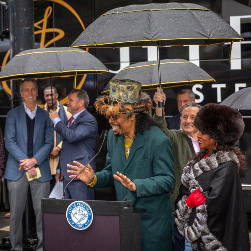 A man in a colorful hat speaks at a podium, surrounded by people holding umbrellas at an outdoor event.