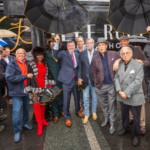A group of people holding umbrellas poses together in front of a building, smiling and dressed in stylish outfits.