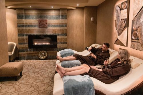 A couple relaxes in spa robes on lounge chairs, enjoying drinks by a cozy fireplace in a serene setting.