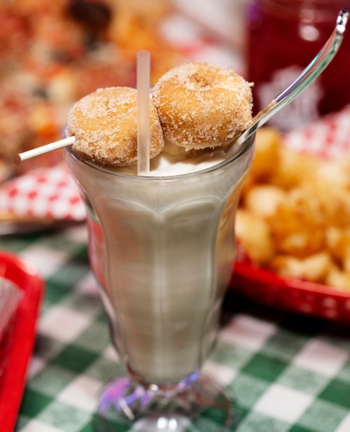 A tall glass of milkshake topped with two mini doughnuts and two straws, set against a checkered tablecloth.
