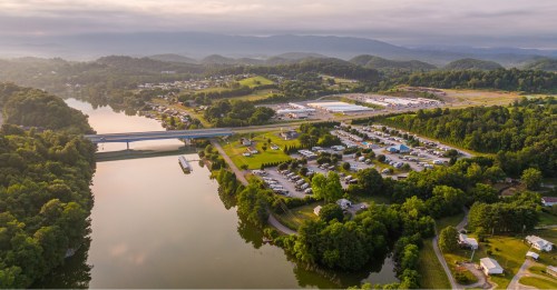 Aerial view of a river, bridge, and green landscape with a small town and mountains in the background.