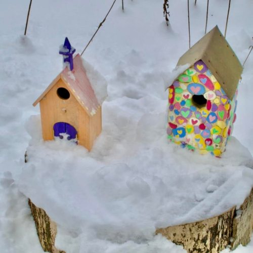 Two colorful birdhouses covered in snow, sitting on a tree stump in a winter landscape.