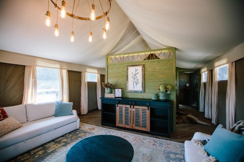 Cozy interior of a tented space featuring a chandelier, sofa, and decorative wall with plants. Natural light from windows.