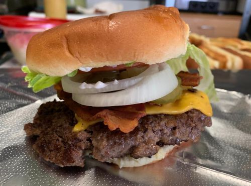 A close-up of a burger with a beef patty, cheese, lettuce, onions, and bacon on a soft bun, resting on foil.