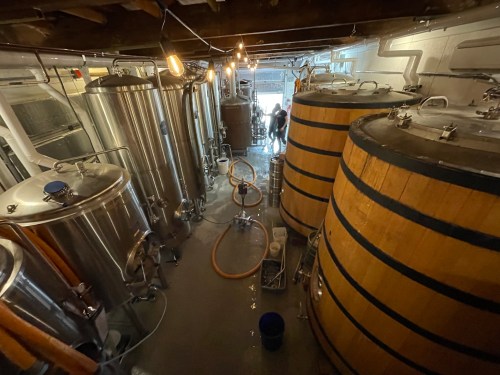Interior of a brewery featuring large metal tanks and wooden barrels, with workers in the background.
