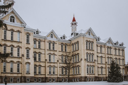 Historic brick building with a red-topped tower, surrounded by snow-covered ground and trees.