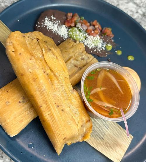 A plate featuring three tamales, black beans, salsa, and a small cup of sauce, garnished with herbs.