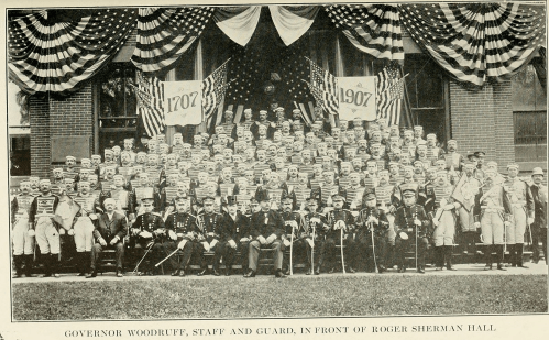 A large group of people in military uniforms and formal attire poses in front of a building decorated with flags and banners.