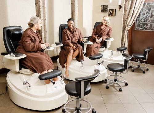 Three women in brown robes relax in spa chairs, enjoying a pedicure in a serene salon setting.