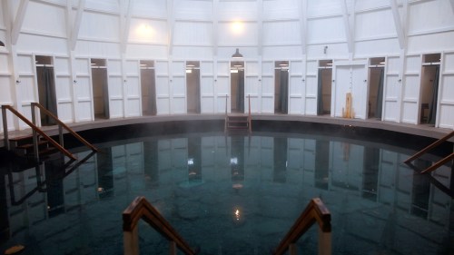 Interior of a circular spa with a central pool, surrounded by wooden steps and private changing rooms. Steam rises gently.