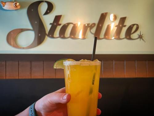 A hand holds a tall glass of yellow drink with ice and a lime slice in front of a "Starlite" sign.