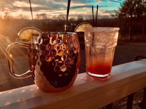 Two cocktails on a railing at sunset, one in a metallic mug with lime slices, the other in a glass with ice.