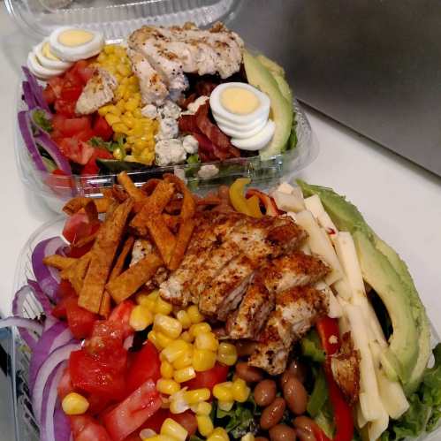 Two colorful salads with grilled chicken, avocado, corn, tomatoes, cheese, and crispy tortilla strips in clear containers.