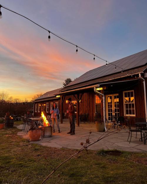 A cozy outdoor setting at sunset, with two people by a fire and string lights illuminating a rustic building.