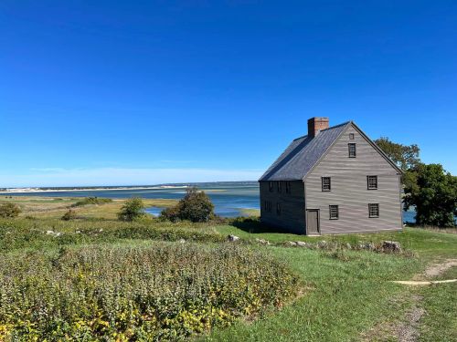 A gray house stands near a calm body of water, surrounded by green fields and clear blue skies.