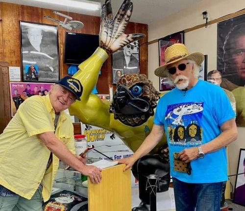 Two men pose beside a large, colorful statue of a superhero in a memorabilia shop, surrounded by framed photos.