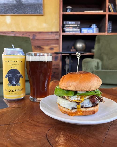 A juicy burger with lettuce, cheese, and a sunny-side-up egg, served with a can of beer and a glass of dark beverage.