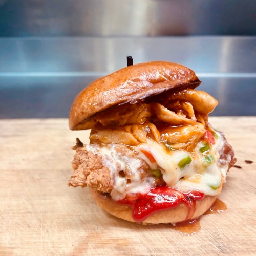 A close-up of a delicious sandwich with fried chicken, melted cheese, and colorful toppings on a toasted bun.