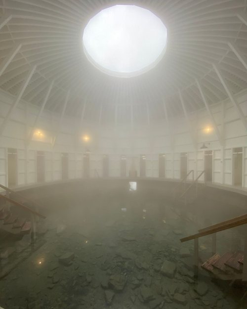 A misty, circular spa interior with a skylight, featuring a pool and wooden steps in a serene atmosphere.