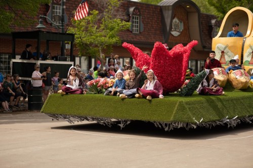 A colorful parade float adorned with flowers, featuring children sitting on a grassy surface, with spectators in the background.