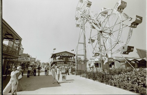 A vintage fair scene featuring a large Ferris wheel, bustling crowds, and amusement stalls under a clear sky.