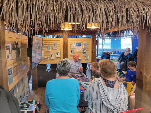 A group of people seated in a cozy, thatched-roof space, listening to a speaker surrounded by informational displays.