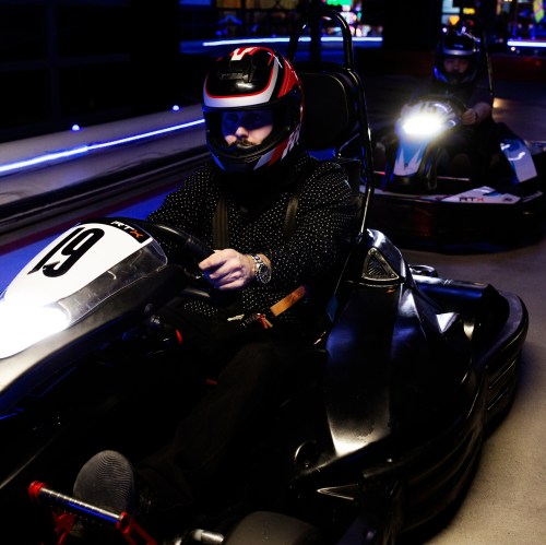 Two racers in go-karts on a track, wearing helmets and racing gear, with colorful lights in the background.