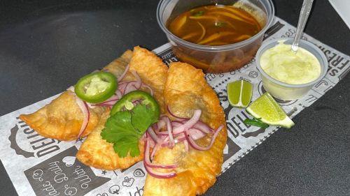 Three crispy empanadas topped with jalapeños and onions, served with a side of soup, lime wedges, and creamy sauce.