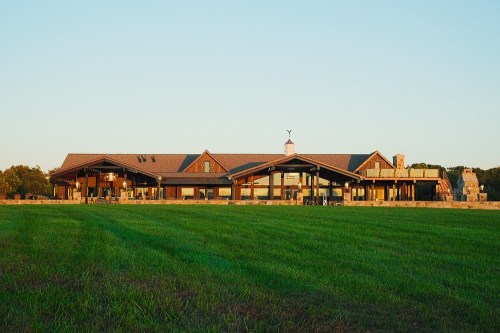 A large, modern building with a sloped roof, surrounded by green grass and trees under a clear blue sky.