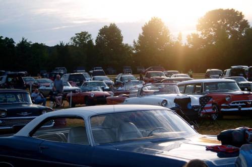 A gathering of classic cars at sunset, with people sitting in lawn chairs among the vehicles.