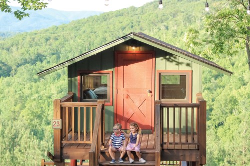 Two children sit on the steps of a colorful treehouse surrounded by lush green mountains.
