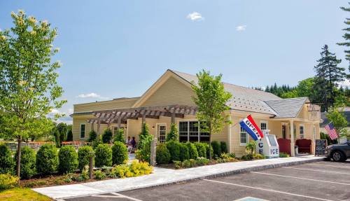 A charming yellow building with a landscaped yard, featuring signs for ice and flags, under a clear blue sky.