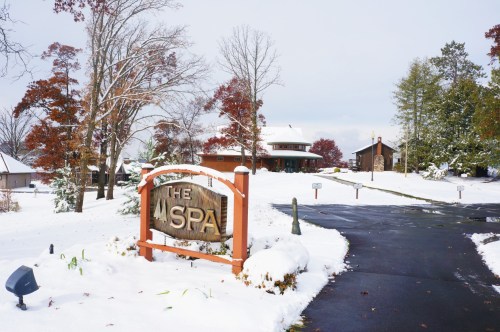 A snowy landscape featuring a sign for "The Spa" with trees and buildings in the background.