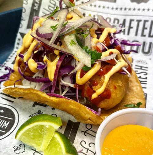 A colorful taco topped with cabbage, onions, and sauce, served with lime wedges and a small cup of orange sauce.