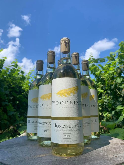 Five bottles of Woodbine Vineyards Honeysuckle Virginia Sweet White Wine against a blue sky and green vineyard backdrop.