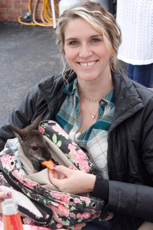 A smiling woman holds a baby kangaroo in a floral bag, feeding it a snack.