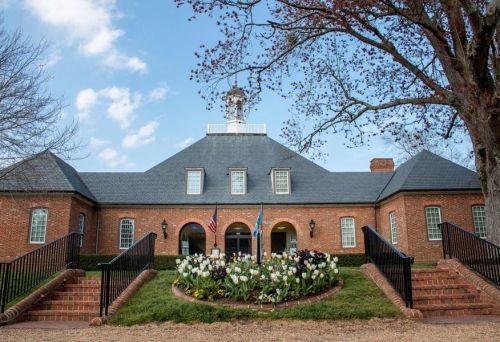 A brick building with a slate roof, flags, and flower beds, set against a blue sky with scattered clouds.