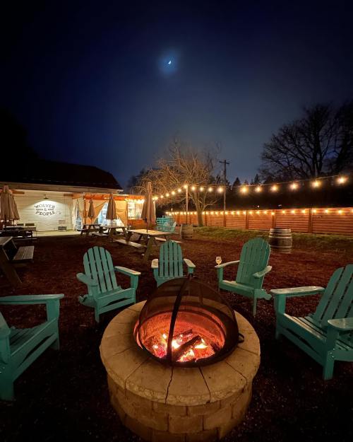 A cozy outdoor space with teal chairs around a fire pit, string lights overhead, and a crescent moon in the night sky.