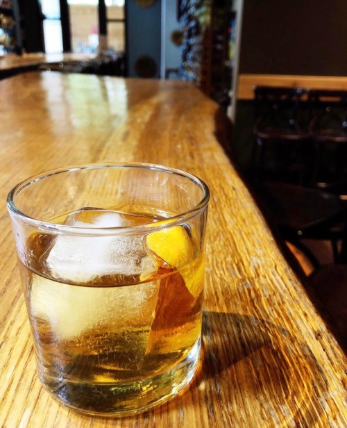 A glass of whiskey with ice and a twist of orange sits on a wooden bar counter.